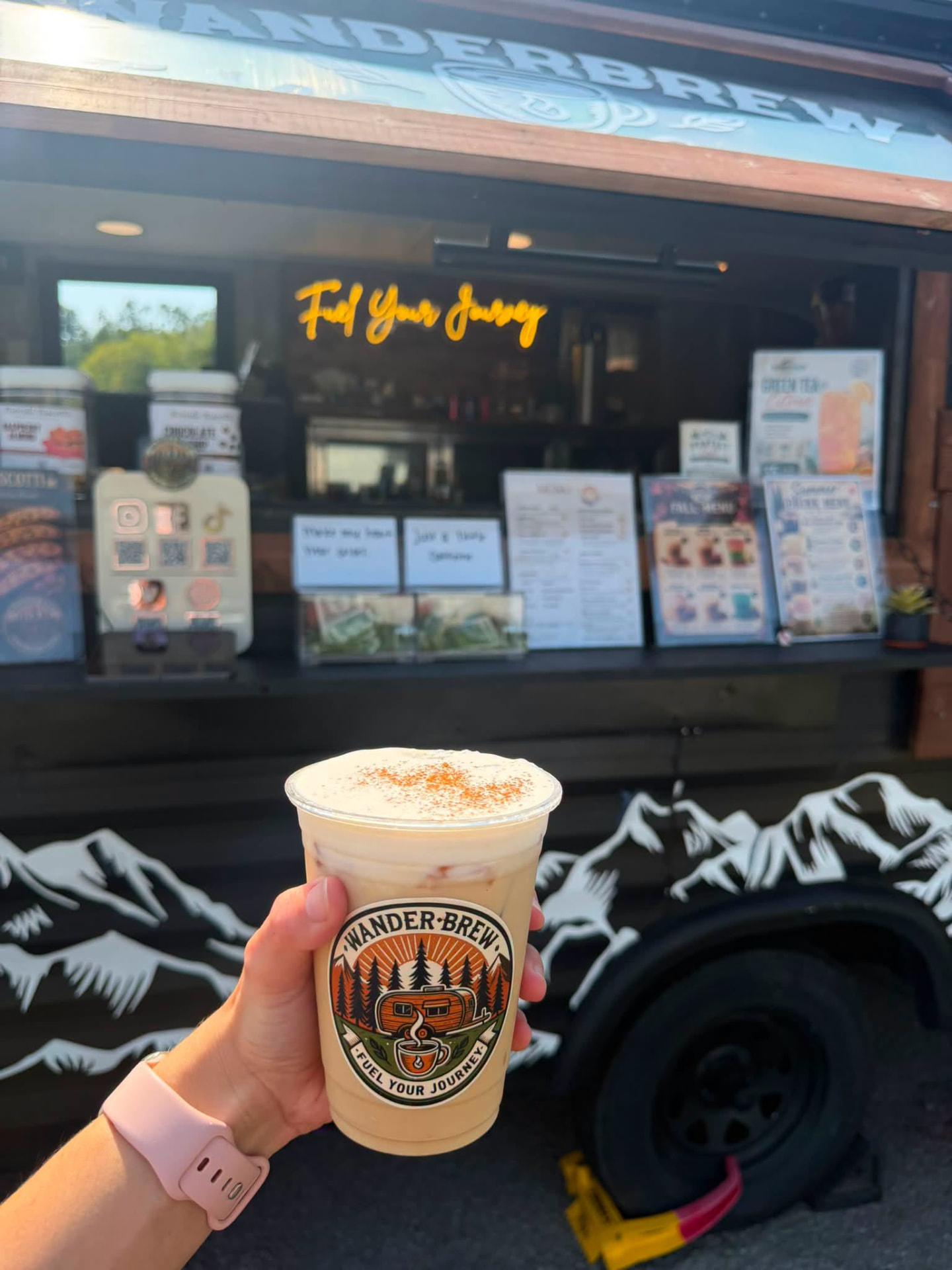 A hand holding an iced coffee topped with cold foam in front of the WanderBrew trailer's service window